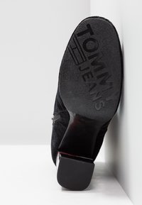 Bottine en daim noir avec semelle extérieure texturée, arborant un logo "TOMMY JEANS" en relief et un talon brillant légèrement incurvé.
