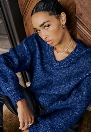 Blauer Strickpullover mit V-Ausschnitt und gerippten Bündchen, der ein strukturiertes Gewebe aufweist. Das Model hat eine silberne Halskette und Ringe als Accessoires.