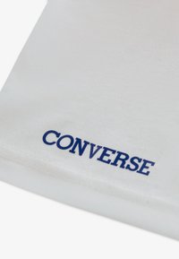 Λευκό ύφασμα με ομαλή υφή που φέρει μπλε εκτύπωση «CONVERSE» κοντά στην κάτω άκρη. Δεν υπάρχουν ορατά μοτί βα ή επιπλέον λεπτομέρειες.