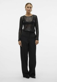 Schwarz, mit Pailletten besetzte Bluse mit langen Ärmeln und einem durchsichtigen Rautenmuster, kombiniert mit hoch geschnittenen, weiten schwarzen Hosen und spitzen schwarzen Schuhen.