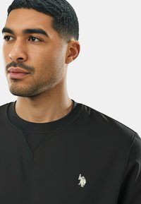 Felpa nera con collo a coste, caratterizzata da un piccolo logo metallico sul lato sinistro del petto e tessuto liscio e morbido.