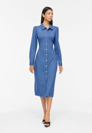Frau trägt ein eng anliegendes langärmliges blaues Midi-Kleid mit Knöpfen und Kragen und geht vor weißem Hintergrund nach vorne.