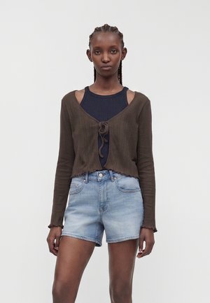 Donna con capelli intrecciati che indossa un cardigan marrone scuro annodato davanti sopra un top a coste blu navy e pantaloncini di jeans azzurri su uno sfondo semplice.