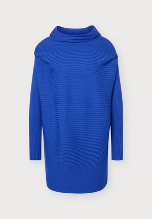 Femme debout, de face, portant une robe-pull côtelée bleu vif et des bottes noires hauteur genoux sur un fond clair uni.