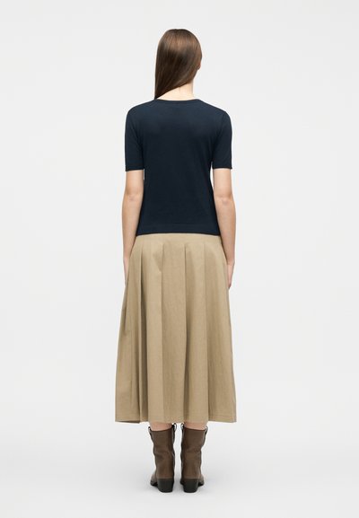 Γυναίκα που στέκεται με την πλάτη, φοράει μπλε σκούρο τοπ με κοντά μανίκια, μπεζ μίντι πλισέ φούστα και καφέ μποτάκια αστραγάλου μπροστά σε απλό φόντο.