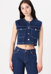 Gilet in denim di colore blu scuro, senza maniche, con bottoni argentati, due tasche frontali e cuciture a contrasto. Indossato con jeans abbinati.