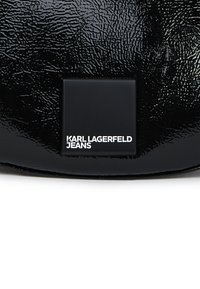 Spīdīgs melns teksturēts materiāls ar mazu matētu melnu kvadrātveida etiķeti, uz kuras baltā tekstā ir ierakstīts "KARL LAGERFELD JEANS".