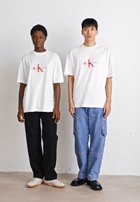 Dva jedinci stojí vedle sebe, mají na sobě bílé grafické trička s logem "Calvin Klein Jeans". Jeden má na sobě černé kalhoty, druhý modré cargo džíny.
