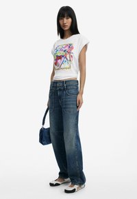 Weißes Grafik-T-Shirt mit buntem Herzdesign, kombiniert mit blauen weiten Jeans und schwarz-weißen Slip-On-Schuhen, hält eine blaue Handtasche.