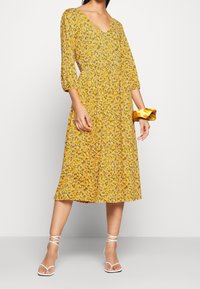 Robe florale jaune avec un décolleté en V et des manches trois-quarts. Le tissu présente des motifs floraux délicats ; assortie à des sandales blanches à lanières et un accessoire jaune.