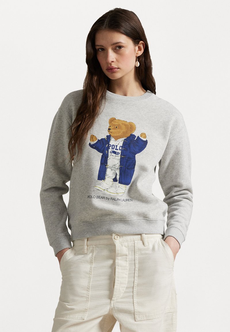 Grauer Sweatshirt mit einem Cartoon-Bären in einer blauen Jacke. Der Bär ist zentriert, mit dem Text "POLO BEAR by RALPH LAUREN" darunter. Weicher Stoff.