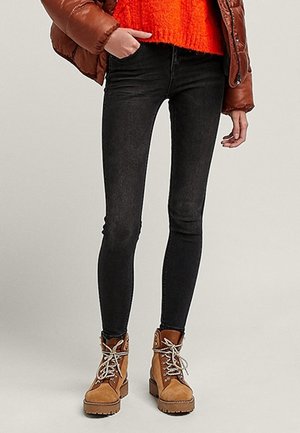 Persoon draagt zwarte skinny jeans, lichtbruine veterlaarzen, feloranje trui en een bruin donsjack met één hand in de zak.