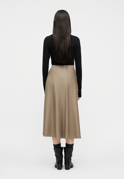 Γυναίκα με μακριά σκουρόχρωμα μαλλιά, ντυμένη με μαύρο μακρυμάνικο τοπ, μπεζ midi φούστα, μαύρες κάλτσες και μαύρα μποτάκια μέχρι τον αστράγαλο, που στέκεται πλάτη προς τα πίσω.