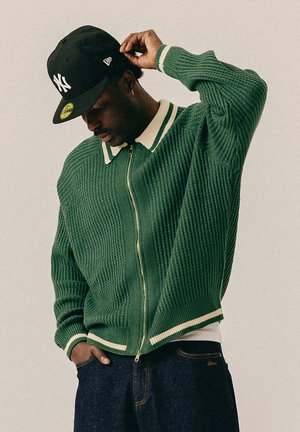 Homme portant un sweat zippé vert avec un bord blanc, un jean foncé et une casquette noire des New York Yankees, ajustant sa casquette d'une main.