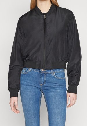 Vrouw draagt een zwarte bomberjas met ritssluiting, geribbelde manchetten en tailleband, gecombineerd met blauwe high-waisted skinny jeans.