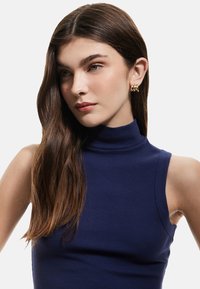 Navy blauwe geribbelde mouwloze coltrui met een slim fit. Het model heeft lang, golvend bruin haar en gouden creolen.