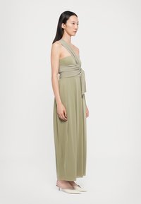 Rochie maxi verde măsliniu, fără bretele, cu un bodice răsucit și un detaliu de legare la talie, realizată dintr-un material fluid. Purtați cu mule cu vârf ascuțit.
