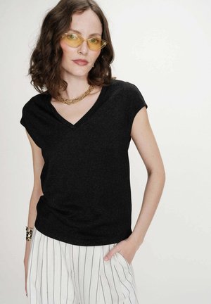 Femme aux cheveux bruns ondulés portant des lunettes de soleil jaunes, un haut noir col en V, un pantalon blanc à rayures noires, un collier chaîne en or et un bracelet.