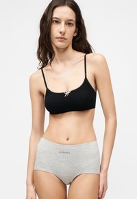 Γυναίκα με μακριά σκούρα μαλλιά που φοράει ένα μαύρο κοντό καμισόλι με λευκό φιόγκο και γκρι εσώρουχα ψηλής μέσης με την επιγραφή "Wednesday".