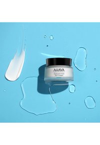 AHAVA Hyaluronsyra 24/7 Cream i en genomskinlig burk med svart lock, placerad på en blå bakgrund med vit kräm och vattendroppar.