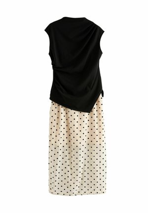Kvinde med krøllet blondt hår iført sort ærmeløs top, beige maxinederdel med prikker, sort clutch og brune sandaler med hæl.