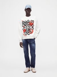 Homme portant un sweat-shirt blanc avec un texte abstrait noir et rouge, un jean bleu, des baskets beige et une casquette bleu clair sur fond blanc.