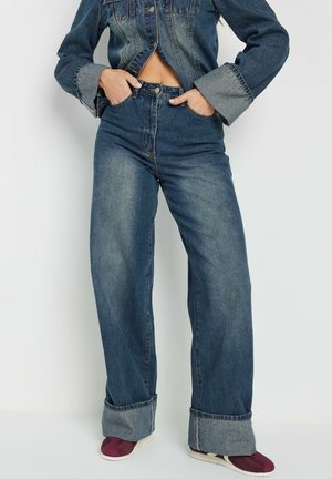 Jeans de cintura alta y pierna ancha en denim azul oscuro con un ligero desgaste. Presenta grandes puños y una cremallera frontal con cierre de botón.