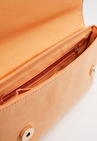 Persikofärgad tygkuvert med en slät yta, utrustad med en zip-invändig ficka och en guldtonad knappförslutning. Fodrad inredning med mjuk finish.