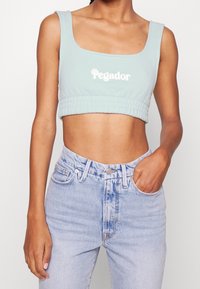 Jasnoniebieski krótki top z haftowanym napisem "Pegador", posiadający kwadratowy dekolt i marszczony dół, zestawiony z jasnoniebieskimi dżinsami.