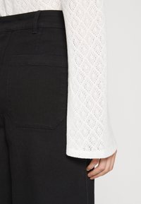 Detailní záběr na osobu v černých kalhotách s zadní kapsou a bílém tričku s dlouhým rukávem a krajkovým vzorem ve tvaru diamantu.