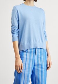 Maglione a maglia di colore azzurro chiaro con maniche lunghe e polsini a coste, caratterizzato da un piccolo logo sul petto, abbinato a pantaloni blu a righe.