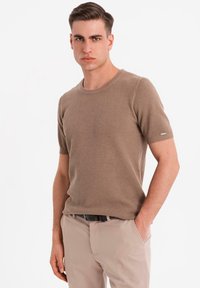 Camisa de punto beige de manga corta con cuello redondo, tela texturizada y un pequeño acento metálico en la manga izquierda, combinada con pantalones claros.