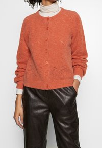 Cardigan en tricot couleur terre cuite avec un col rond, des manches longues et une fermeture à boutons. Présente un motif texturé et des accents de boutons contrastés.