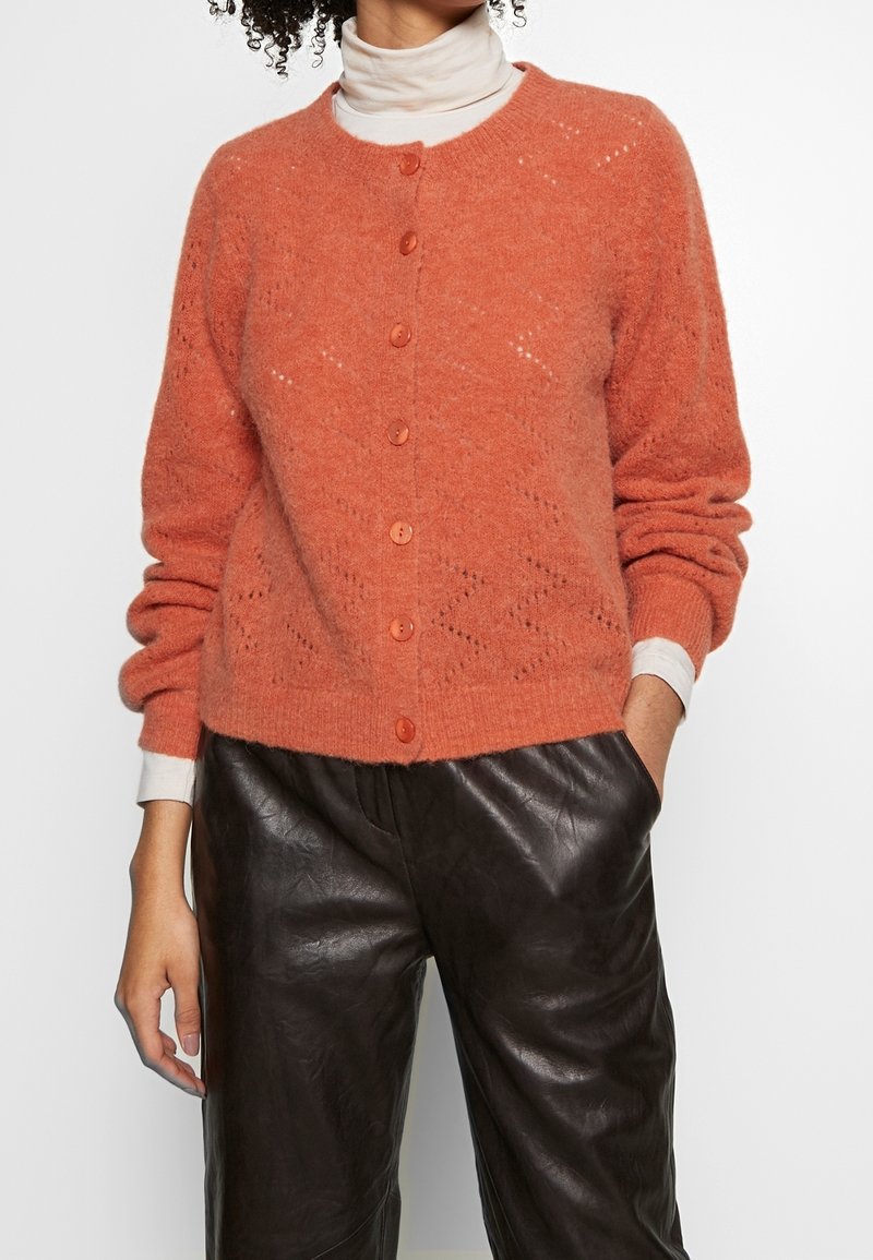 Cardigan en tricot couleur terre cuite avec un col rond, des manches longues et une fermeture à boutons. Présente un motif texturé et des accents de boutons contrastés.