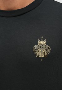 Czarna koszulka z logo w kolorze złotym przedstawiającym psa i ozdobny wzór. Okrągły dekolt i krótkie rękawy wykonane z gładkiej bawełny.