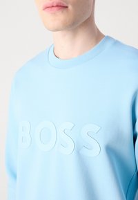Közelkép egy személyről, aki egy világoskék pulóvert visel, amelynek mellrészén emelt betűk formájában a "BOSS" felirat olvasható.