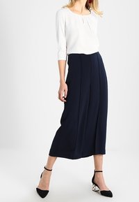 Marineblaues Culotte mit hoher Taille und Falten, kombiniert mit einer weißen Bluse, die ein Schlüsselaugen-Detail aufweist. Schwarze und weiße spitze Absatzschuhe sind im Lieferumfang enthalten.