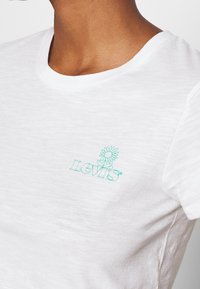 Bílý bavlněný tričko s kulatým výstřihem a krátkými rukávy, se zeleným květinovým logem Levi's na levé části hrudníku.