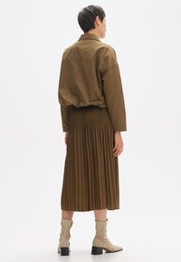 Veste cropped marron sur une jupe plissée marron. Le mannequin porte des bottines beige à talon carré pour une apparence superposée et texturée.