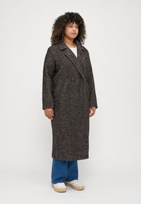 Brun herringbone-mønstret lang frakk med dobbel knappestolpe, bred krage, to sidelommer og en avslappet passform.