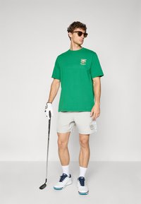 Mann iført grønn t-skjorte, lyse shorts, golfhanske, solbriller og hvite golf sko, som holder en golfkølle og står foran en ensfarget bakgrunn.
