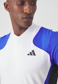 Korte mouwen sporttop gemaakt van gladde, vochtafvoerende stof. Heeft blauwe schouders, een witte romp en een zwart logo op de borst.