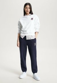 Felpa bianca in pile con logo, indossata sopra una camicia a fantasia chiara. Pantaloni della tuta blu navy con caviglie elasticizzate e dettagli con logo. Sneakers bianche.