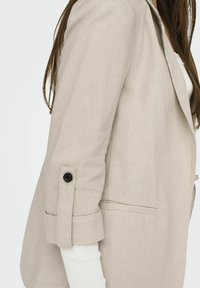 Blazer in lino beige con rever a punta, maniche arrotolate con bottoni neri e taglio dritto. Sotto si intravede una camicia bianca a maniche lunghe.