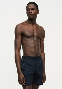 Μυώδης άνδρας που στέκεται μπροστά σε απλό φόντο, φορώντας σκούρα μπλε μαγιό με κορδόνι στη μέση, κοιτάζοντας ελαφρώς προς τα πλάγια.