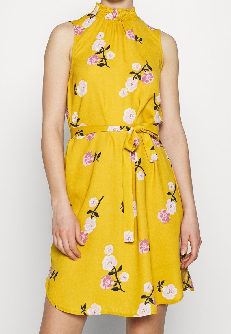 Robe fleurie jaune avec un col montant, sans manches, et une ceinture à nouer à la taille. Présente des motifs de fleurs roses et blanches sur un tissu texturé.