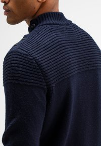 Marine gebreide trui met een ribbelkraag en schouderpatroon, voorzien van drie knopen bij de hals. Zachte textuur en slim fit ontwerp.