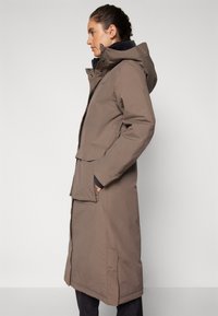 Long brown waterproof parka with a hood, featuring a zip and snap closure, side pockets, and a streamlined silhouette.