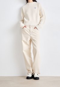 Pull crème côtelé avec un col rond, associé à un pantalon large blanc cassé avec des surpiqûres contrastantes et des chaussures noires plates.