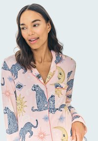 Mönstrad pyjamas överdel i mjuk rosa tyg, prydd med lejon, solar och ögon. Anmärkningsvärda detaljer inkluderar kontrastsömmar och en ficka.